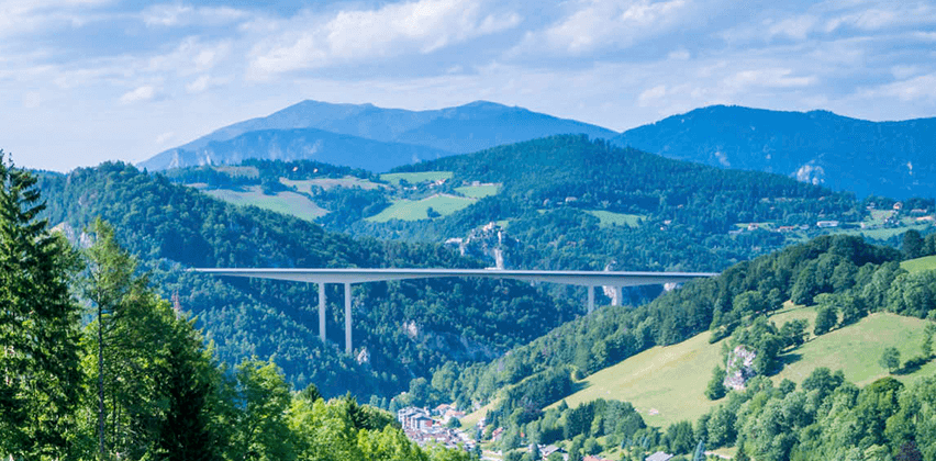 Ein Blick von einem bewaldeten Hang auf eine lange Brücke, die sich über ein Tal mit Bergen im Hintergrund erstreckt.