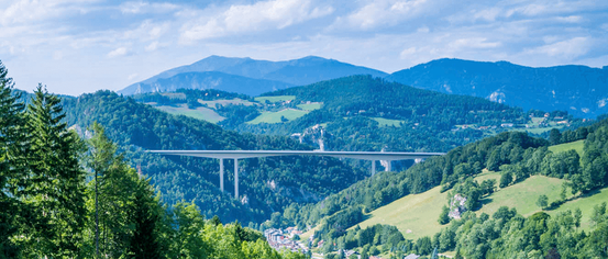 Ein Blick von einem bewaldeten Hang auf eine lange Brücke, die sich über ein Tal mit Bergen im Hintergrund erstreckt.