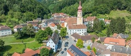 Ein Luftbild eines kleinen Ortes mit einem Kirchturm in der Mitte, umgeben von Häusern, Bäumen und Grünflächen. Der Ort liegt in einem Tal mit klarem blauem Himmel.
