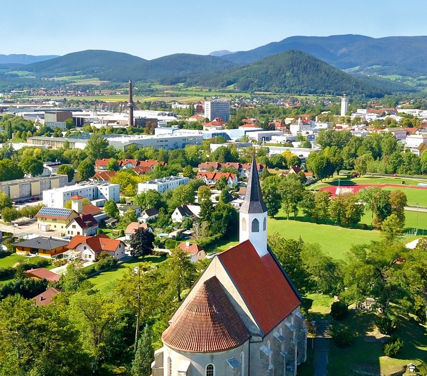 Ein Luftbild einer Stadt mit Häusern und einer Kirche mit rotem Dach. Die Stadt ist von Bäumen und einem Rasenfeld umgeben. In der Ferne befinden sich Berge.