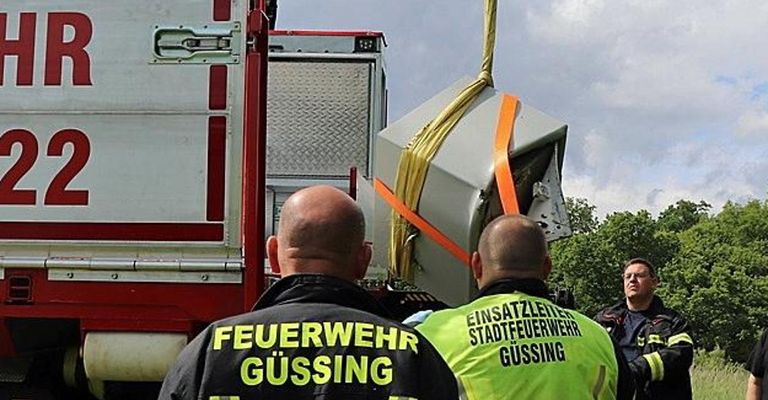 Zwei Feuerwehrleute heben ein beschädigtes Gerät auf einem Feuerwehrwagen an, wobei einer eine Gusing Feuerwehrjacke und der andere eine Gusing Stadtfeuerwehrjacke trägt.