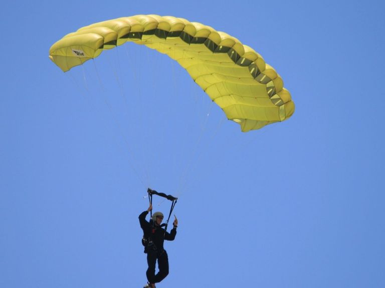 Eine Person ist im Himmel beim Paragliden, trägt einen Helm und Gurt, hält sich an der Handhabe des Fallschirms fest, mit einem gelben Fallschirm gegen einen blauen Himmel.