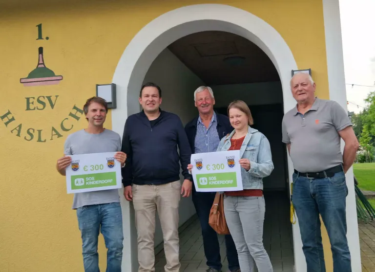 Fünf Personen stehen vor einer gelben Wand mit einem Schild, lächeln und halten Schilder mit 300€ und SOS Kinderdorf. Die Frau rechts trägt eine Jeansjacke und hält eine braune Handtasche.
