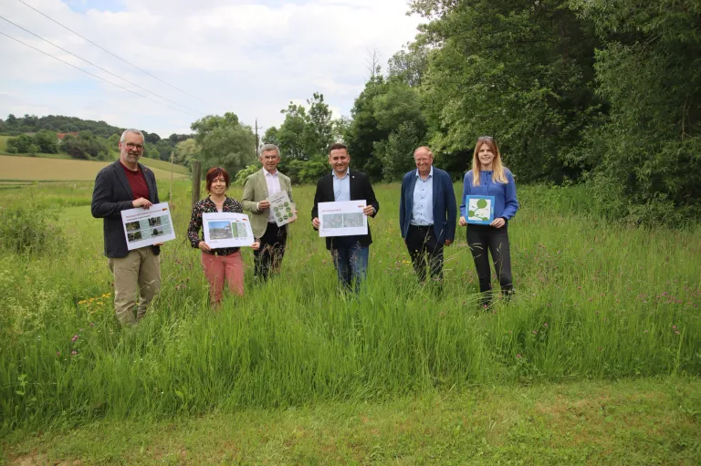 Eine Gruppe von Menschen steht in einem Feld mit hohem Gras und hält weiße Papiere mit grünen Symbolen. Sie scheinen für ein Foto in einer ländlichen Umgebung zu posieren.