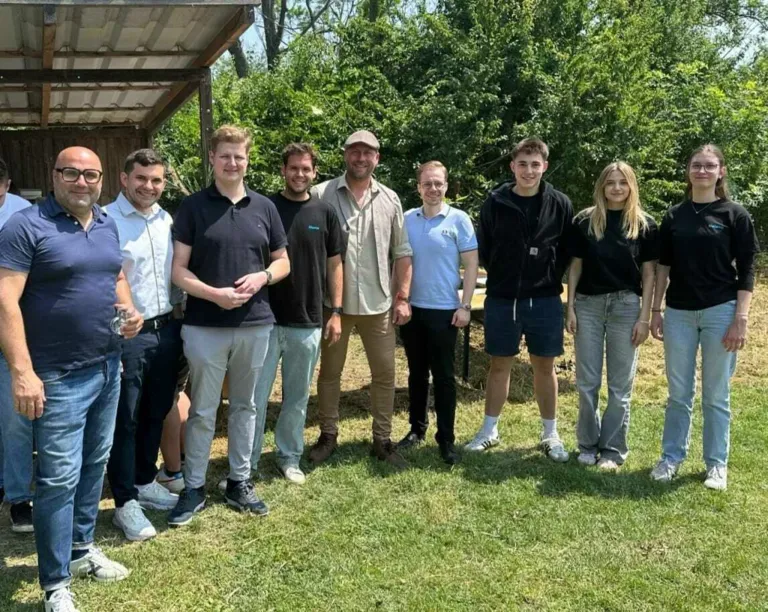 Eine Gruppe von Menschen posiert für ein Foto in einem Garten mit einer Holzstruktur und Bäumen im Hintergrund. Sie tragen Freizeitkleidung.