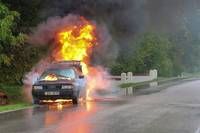 Ein Auto brennt auf einer nassen Straße, mit Rauch, der aus dem Motor aufsteigt und Flammen, die das Dach lecken. Das Auto ist am Straßenrand geparkt.