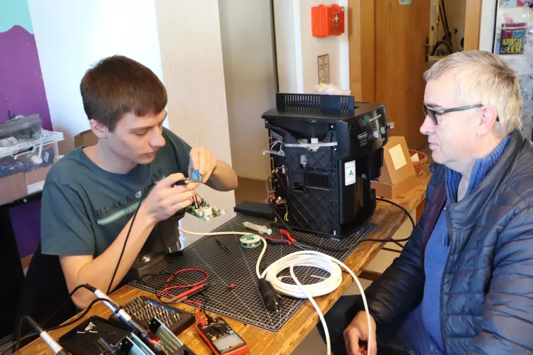 Zwei Männer arbeiten in einer Werkstatt an einer Maschine. Einer justiert Drähte, während der andere beobachtet. Werkzeuge und Kabel liegen auf dem Tisch.