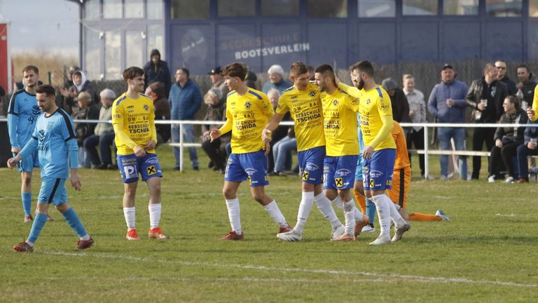 Eine Gruppe von Fußballspielern in gelben und blauen Trikots feiert auf einem Feld. Im Hintergrund ist ein Zaun und Zuschauer zu sehen. Werbung ist am Zaun und Gebäude sichtbar.