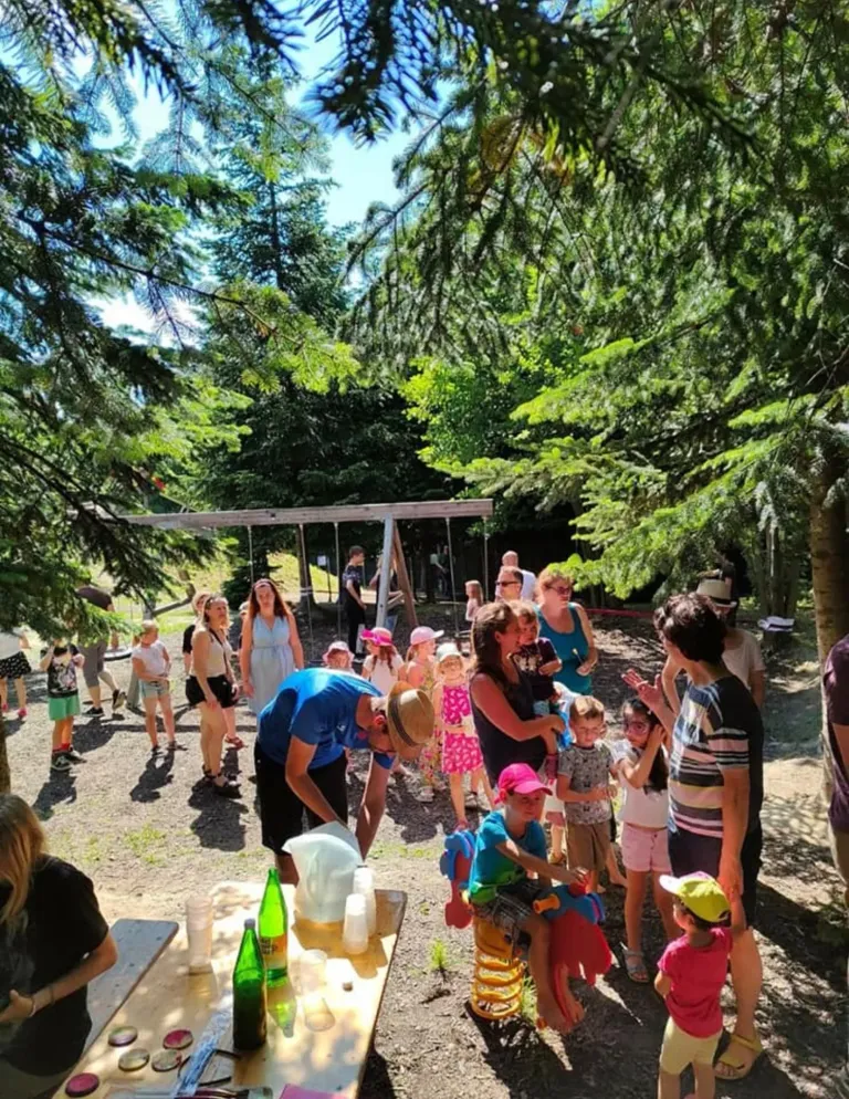 Eine Gruppe von Menschen, einschließlich Erwachsener und Kindern, befindet sich in einem Park. Einige stehen herum, andere sitzen auf Schaukeln, und eine Person gießt etwas in einen Becher.
