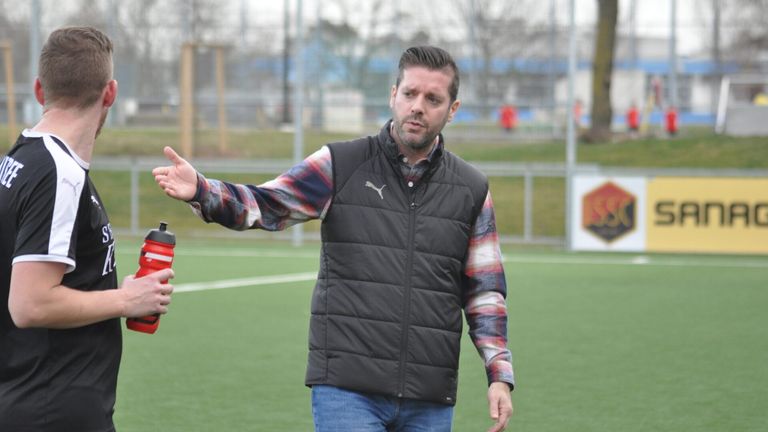Ein bärtiger Mann steht auf einem Fußballfeld und gestikuliert mit der Hand. Er trägt eine schwarze Weste und ein kariertes Hemd.