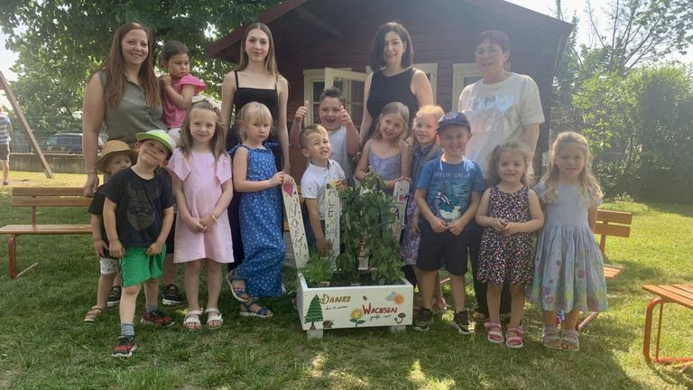 Eine Gruppe von Erwachsenen und Kindern steht lächelnd vor einem Haus für ein Foto. Die Kinder halten Schilder hoch, und eine Topfpflanze steht in der Mitte.