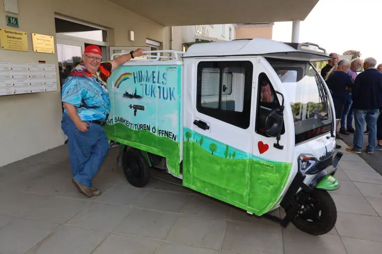 Ein Mann steht neben einem bemalten weißen und grünen Elektrofahrzeug mit 'Himmels Tuktuk' darauf, wobei er sich am Dach festhält.