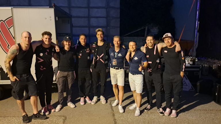 Sieben Menschen stehen in einer Reihe und lächeln für ein Foto. Sie sind in passenden Outfits gekleidet. Hinter ihnen sind ein Fahrzeug und ein Gebäude zu sehen.