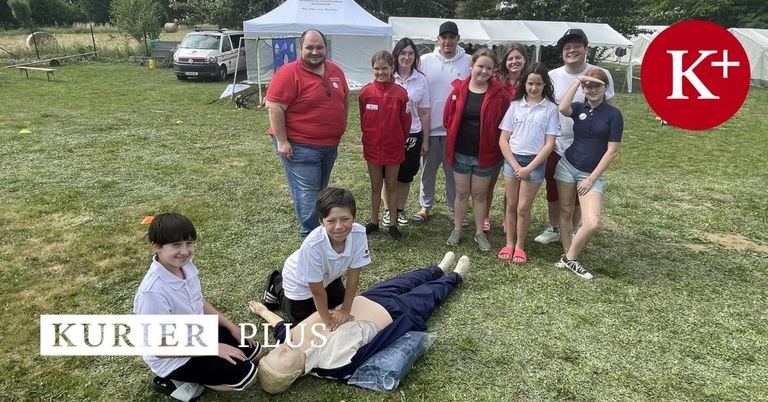 Eine Gruppe von Menschen in passenden roten und weißen Shirts, zusammen mit Erwachsenen, stehen und knien auf einem Rasenfeld mit Zelten im Hintergrund.