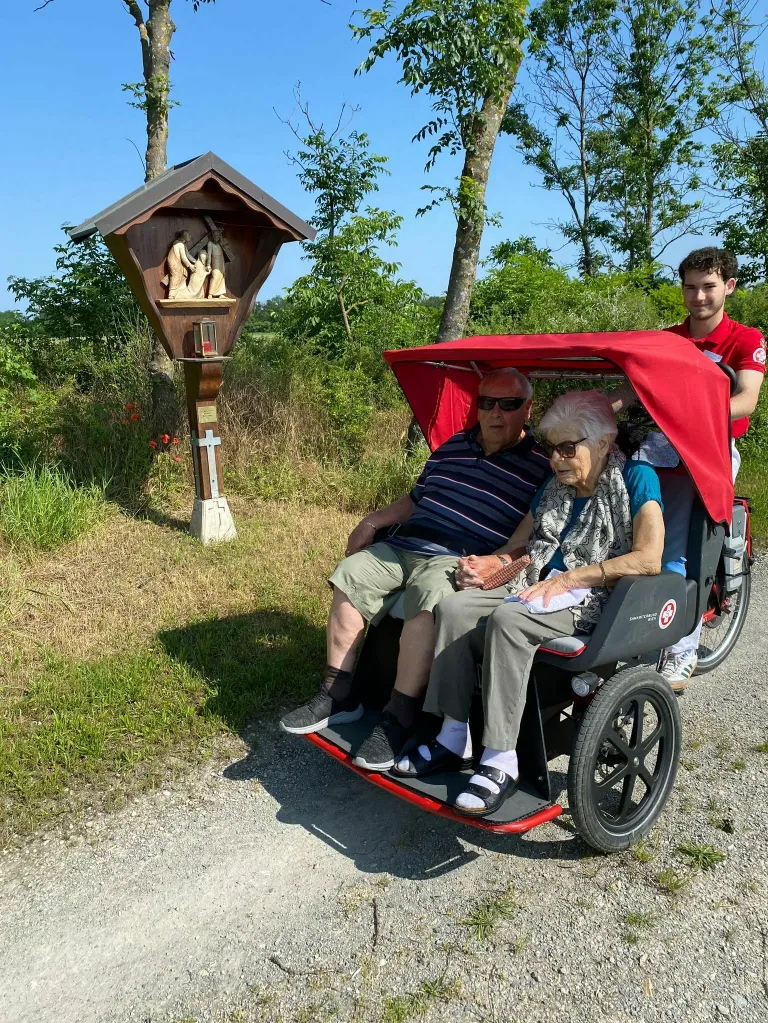 Ein älteres Paar fährt ein Dreirad im Freien, ein junger Mann schiebt es. Dahinter steht ein hölzernes Heiligtum mit einem Kreuz und einer Statue.
