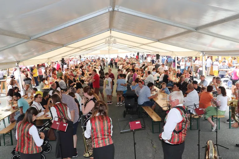 Eine große Menschenmenge versammelt sich unter einem Zelt mit Tischen und Stühlen. Die Leute sitzen und stehen, essen und trinken. Ein Mann spielt Trompete und ein anderer steht vor einem Notenständer. Einige Menschen tragen traditionelle Kleidung.