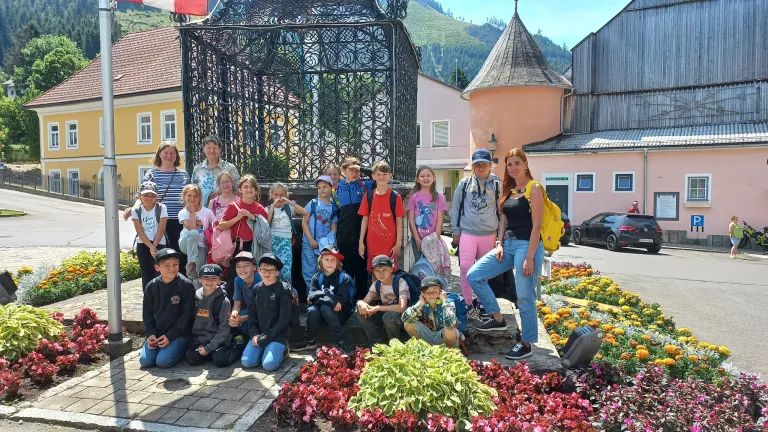 Eine Gruppe von Kindern und Erwachsenen posiert für ein Foto vor einem dekorativen Brunnen, mit Gebäuden und Bergen im Hintergrund.