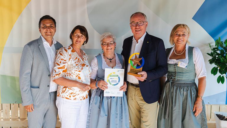 Eine Gruppe von Erwachsenen steht lächelnd zusammen, eine Frau hält ein Zertifikat. Hinter ihnen hält ein Mann einen goldenen Stern-Preis. Sie sind alle formell gekleidet.