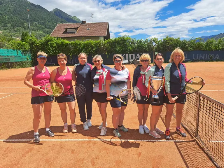 Sieben Frauen posieren auf einem Tennisplatz, halten Tennisschläger und Pokale und haben ein Haus und Berge im Hintergrund.