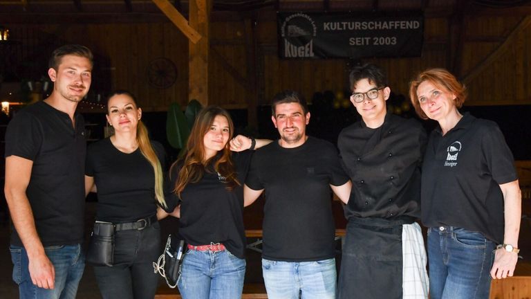 Vier Personen, die in Schwarz gekleidet sind, stehen lächelnd zusammen in einer rustikalen Umgebung. Sie tragen Freizeitkleidung, und ein Banner im Hintergrund zeigt 'KUGEL' und 'SEITE 2003'.