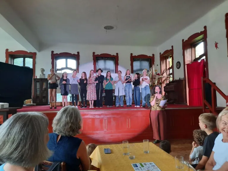 Eine Gruppe von Menschen steht auf der Bühne, einige klatschen. Eine Frau hält ein Mikrofon und spricht zu ihnen. Die Bühne ist mit rotem Stoff geschmückt und ein Klavier steht in der Nähe. Das Publikum sitzt vor einem Tisch mit Gläsern.