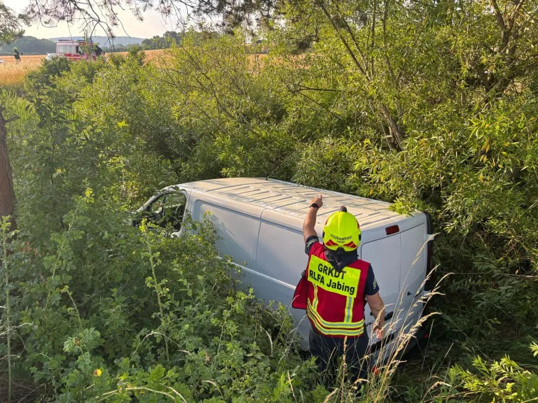 Ein Feuerwehrauto steht im Hintergrund, während ein Feuerwehrmann mit Weste und Helm auf einen Van zeigt, der in dichtes Gebüsch gecrasht ist.