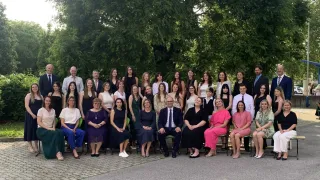 Eine Gruppe von Menschen, darunter Männer und Frauen, posiert für ein Foto im Freien. Sie sitzen auf Stühlen, und einige stehen. Sie sind alle formell gekleidet. Hinter ihnen stehen große Bäume.