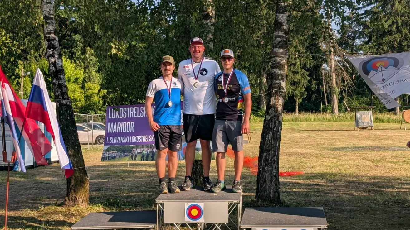 Drei Männer stehen auf einem Podium mit Medaillen um den Hals. Sie tragen Sportkleidung und lächeln für ein Foto. Dahinter befindet sich ein Banner mit dem Text 'LUKOSTRELSI MARIBOR' und einem Logo. Bäume und Gras sind im Hintergrund.