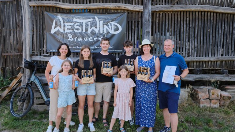 Eine Familie steht vor einer Holzstruktur mit einem Banner, das 'Westwind Brauerei' besagt. Sie halten Bierflaschen in den Händen.