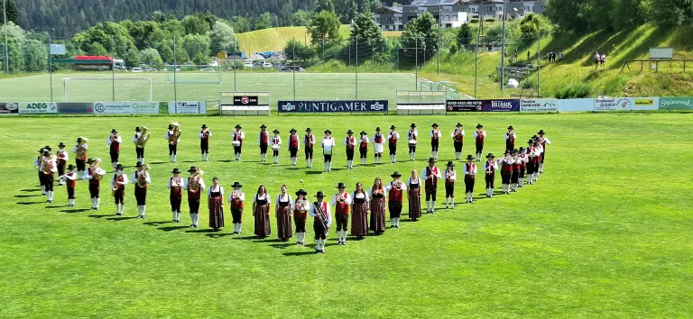 Eine Marschkapelle in traditionellen Kostümen, die auf einem Fußballfeld steht, mit Bannern dahinter. Sie halten Musikinstrumente und scheinen eine Aufführung zu machen.