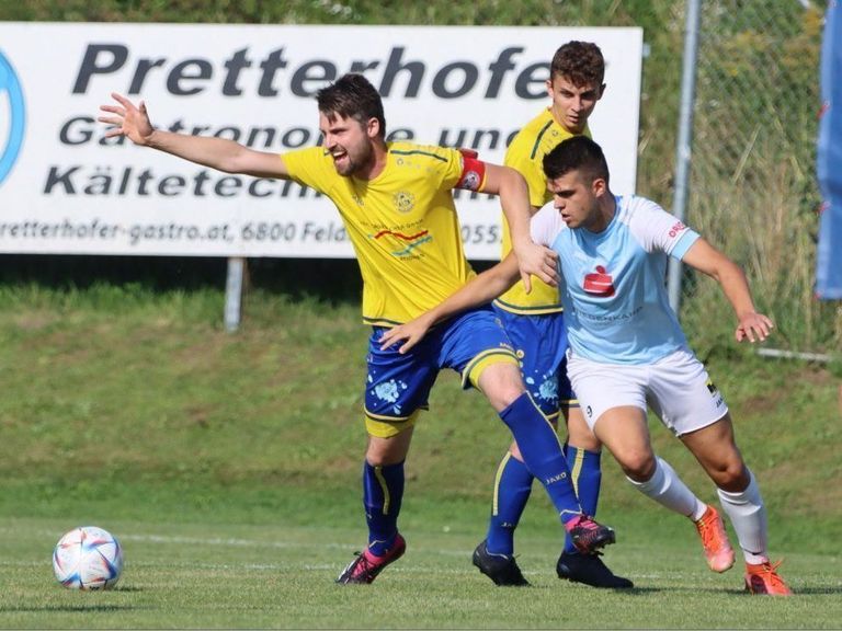 Drei Fußballspieler sind auf einem Rasenfeld in einem Spiel engagiert. Ein Spieler in Gelb streckt seinen Arm aus, während ein anderer in Blau ihn tackelt. Der dritte Spieler in Weiß versucht, den Ball zu bekommen.
