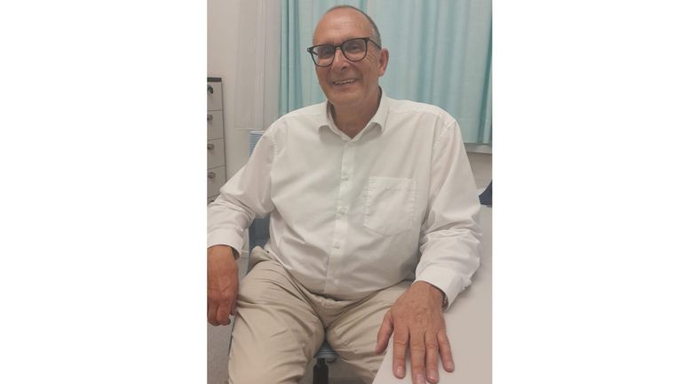 Ein Mann mit einer Brille und einem weißen Langarmhemd sitzt auf einem Stuhl mit einem Lächeln im Gesicht. Er hält einen Stift in der Hand und scheint für ein Foto zu posieren. Hinter ihm ist eine weiße Wand mit einem blauen Vorhang.