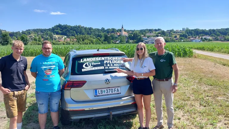 Drei Personen stehen vor einem silbernen Auto. Ein Mädchen berührt das Auto und lächelt. Ein Mann in einem grünen Hemd posiert mit ihr. Das Auto hat ein Schild mit den Worten Landesentscheid Pflugen.