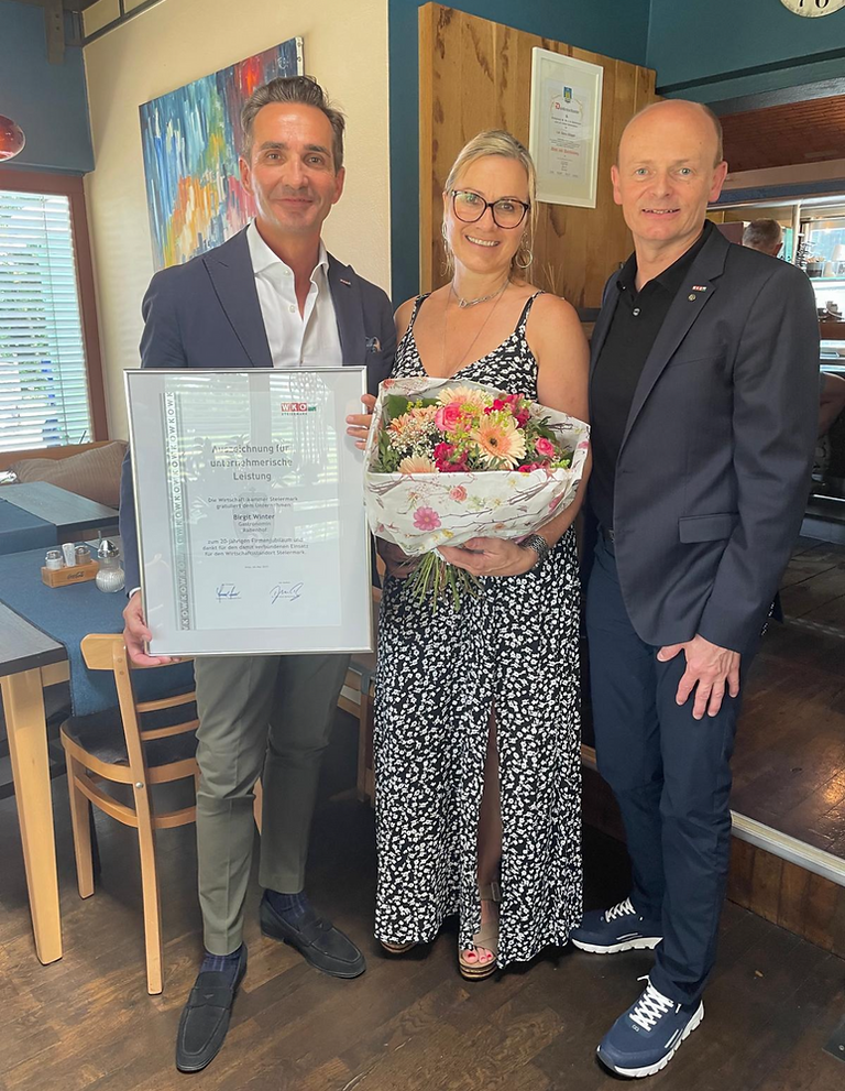 Drei Personen, zwei Männer und eine Frau, stehen in einem Restaurant. Die Frau hält einen Blumenstrauß und ein Zertifikat. Die Männer tragen Anzüge, und einer hält ein gerahmtes Zertifikat. Hinter ihnen sind Tische und Stühle zu sehen.