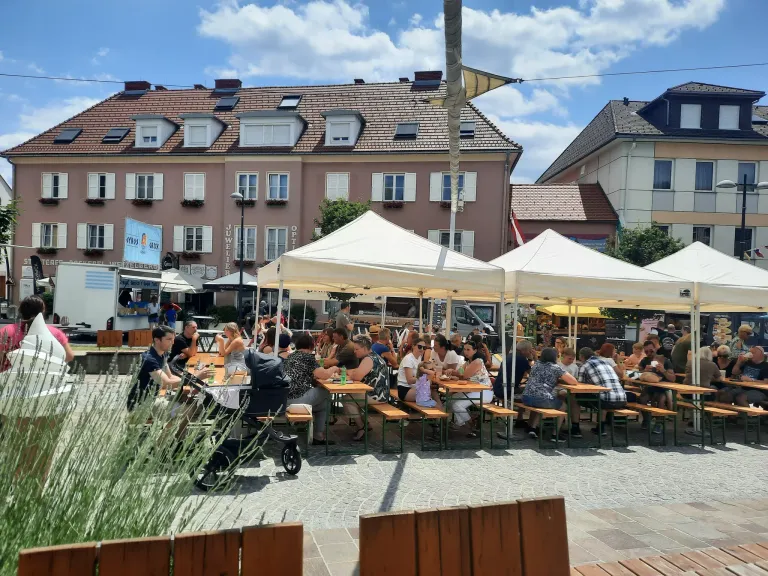 Terrasse eines belebten Cafés mit vielen Menschen, die das Sommerwetter unter weißen Zelten genießen, mit Gebäuden im Hintergrund.