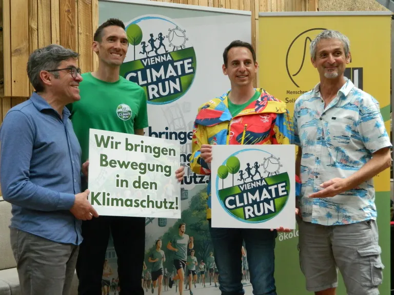 Vier Männer stehen vor einem Plakat, lächeln und halten Schilder zur Förderung des Vienna Climate Run. Der Mann links trägt ein grünes T-Shirt mit Logo, der Mann in der Mitte ein farbenfrohes Jackett. Der Mann rechts trägt ein gemustertes Hemd.