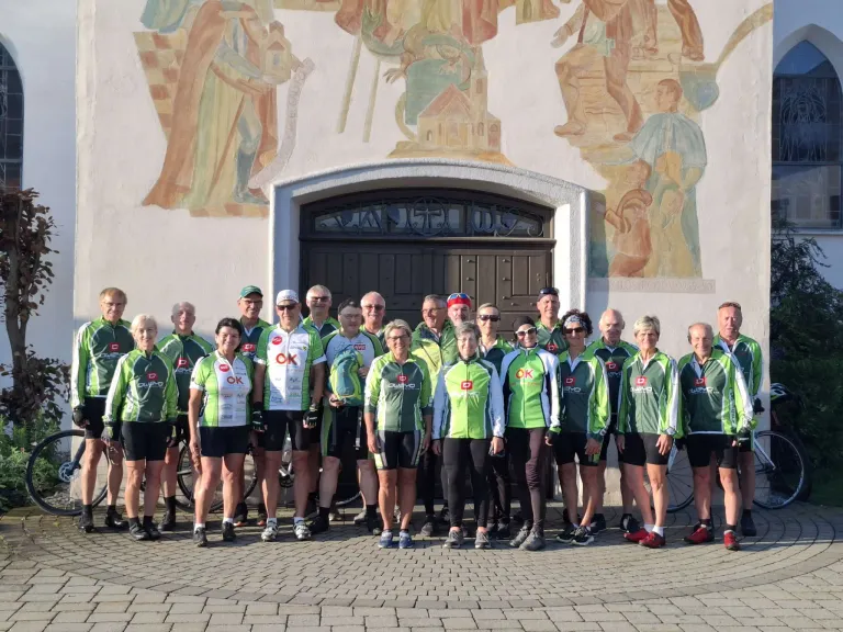 Eine Gruppe von Menschen in grüner Radsportausrüstung steht vor einem Gebäude mit einem Wandbild. Sie lächeln und posieren für ein Gruppenfoto.