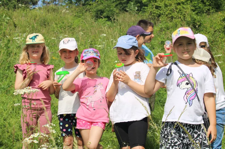 Eine Gruppe von Kindern in einem Grasfeld, die kleine Behälter mit Flüssigkeit darin halten. Sie scheinen an einem wissenschaftlichen Experiment beteiligt zu sein. Sie tragen Mützen und Brillen.