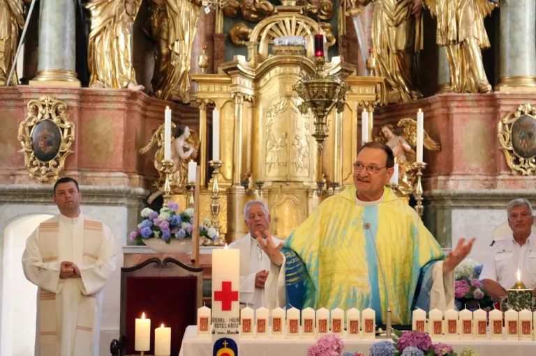 Ein Priester in einer gelben und blauen Robe steht vor einem goldenen Altar mit Kerzen und Blumen. Er lächelt und scheint zu sprechen. Hinter ihm schaut ein anderer Priester zu.