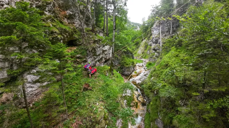 Eine Person mit einem roten Rucksack sitzt auf einem felsigen Hang und bereitet sich auf eine Seilabfahrt vor. Grüne Vegetation und Bäume umgeben das Gebiet. Ein kleiner Bach fließt durch die Mitte des Berges.