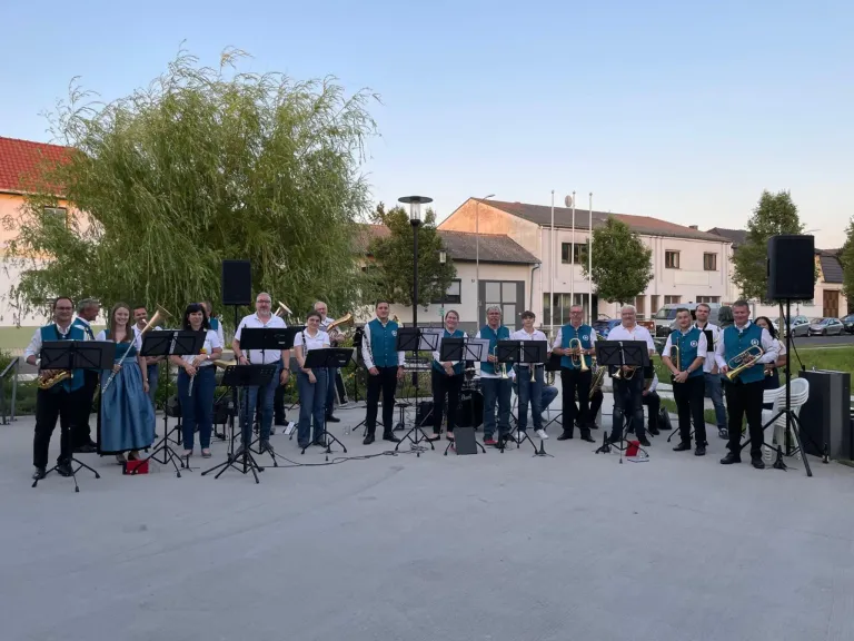 Eine Band spielt vor einem Gebäude mit mehreren Fenstern. Sie stehen vor Notenständern mit verschiedenen Instrumenten.
