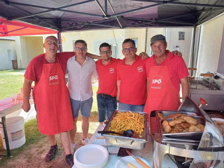 Fünf Männer in roten Hemden und Schürzen stehen unter einem Zelt. Sie lächeln und posieren für ein Foto. Vor ihnen steht ein Tisch mit Essen auf Tabletts.