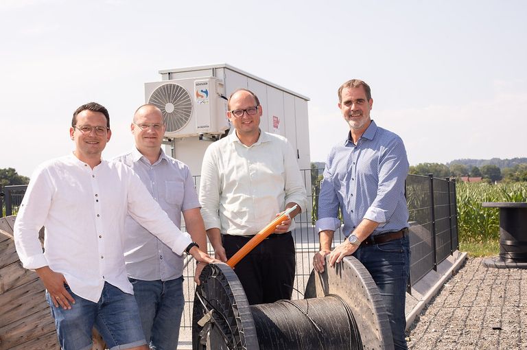 Vier Männer stehen draußen in der Nähe eines Zauns und lächeln für ein Foto. Einer hält eine große orange Spule Kabel. Dahinter steht eine weiße Struktur mit einer Klimaanlage.