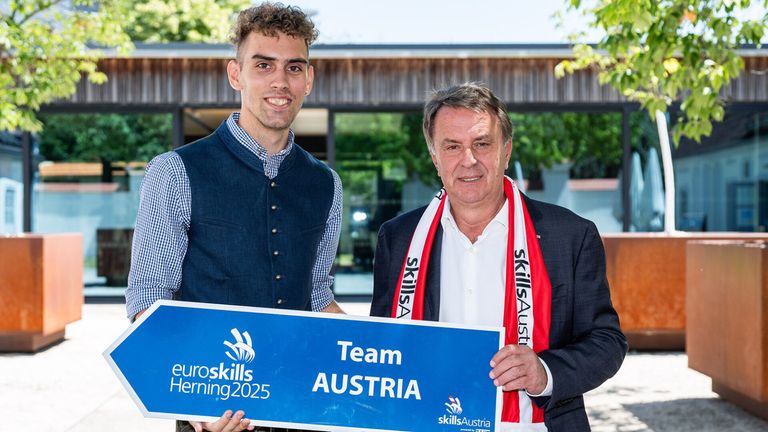 Zwei Männer stehen zusammen und halten ein blaues Schild, auf dem 'Team AUSTRIA' mit dem 'euroskills Herning 2025'-Logo steht. Der Mann auf der linken Seite lächelt, während der Mann auf der rechten Seite einen rot-weißen Schal trägt. Sie stehen vor einem Gebäude mit Glasfenstern.