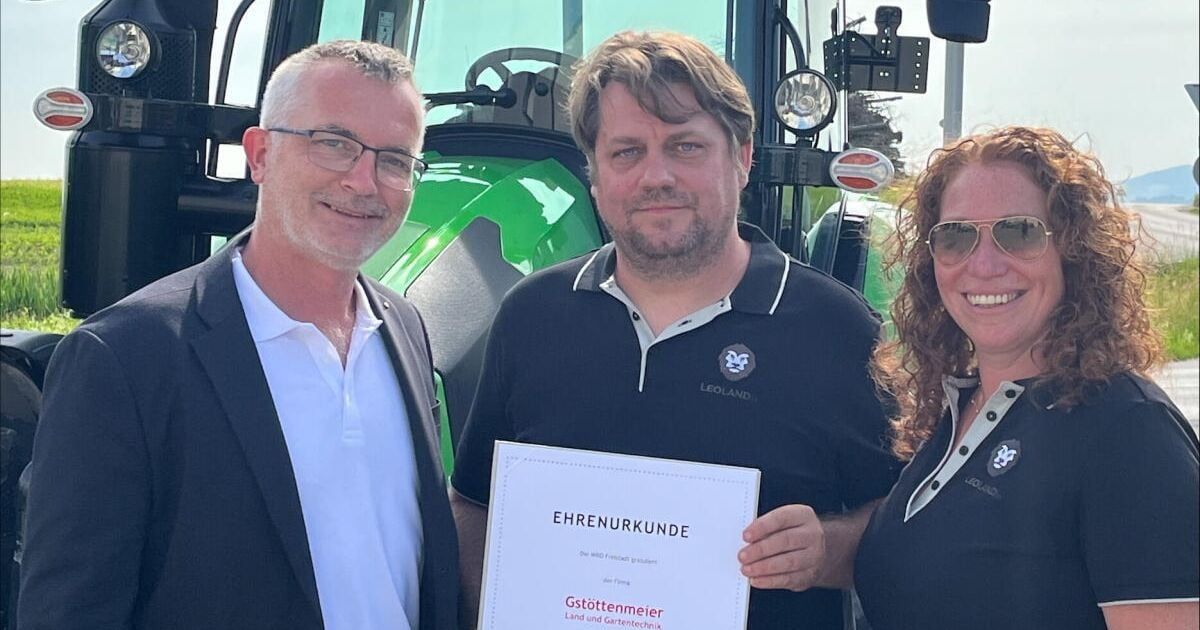 Drei Personen stehen vor einem grünen Traktor. Ein Mann in der Mitte hält ein Zertifikat mit der Aufschrift 'Ehrenurkunde'. Der Mann links trägt einen Anzug und die Frau rechts trägt eine Sonnenbrille.