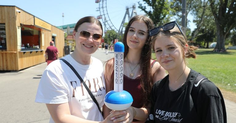 Drei Frauen posieren mit einem Thermometer in einem Freiluft-Vergnügungspark, mit einem Riesenrad im Hintergrund.