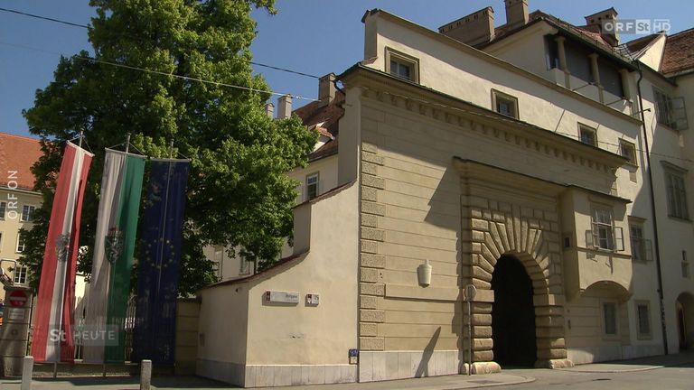 Ein Gebäude mit beigefarbenen Wänden und gewölbten Torbögen an einem sonnigen Tag. Vor dem Gebäude steht ein Baum und ein Schild ist an der Wand befestigt.
