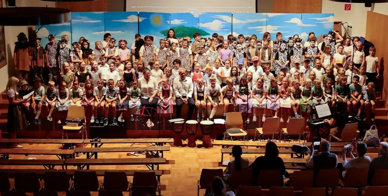 Eine Gruppe von Kindern und Erwachsenen in verschiedenen Kostümen sitzt auf der Bühne, möglicherweise bei einer Aufführung. Trommeln und ein Mikrofon stehen auf der Bühne. Dahinter ist eine bemalte Kulisse mit einer Sonne und einem Baum. Einige Menschen sitzen vor der Bühne auf Stühlen.