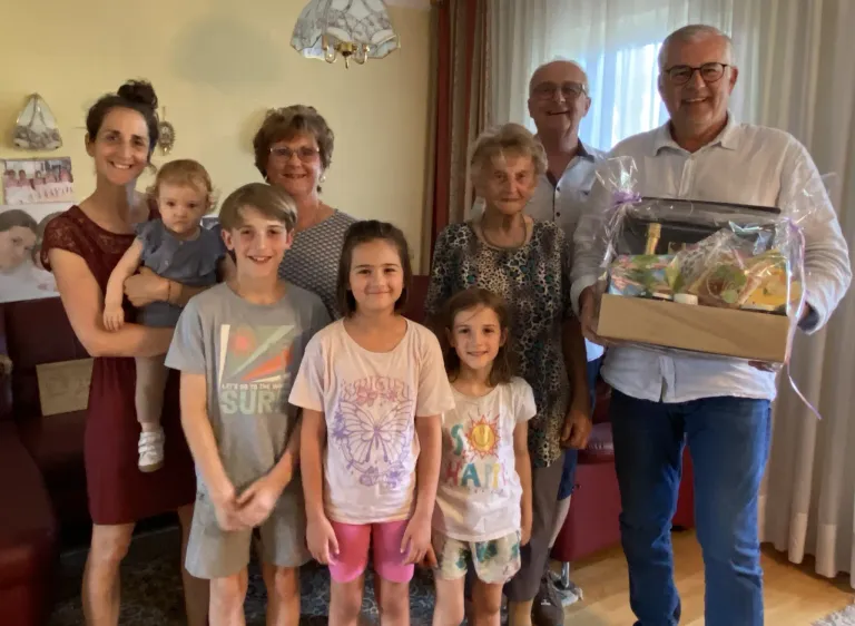 Ein Familientreffen im Wohnzimmer. Zwei ältere Frauen und ein Mann lächeln. Ein Mann hält eine Geschenkbox. Kinder und ein Baby sind ebenfalls anwesend.