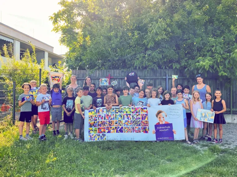 Eine Gruppe von Kindern und Erwachsenen steht in einem Garten mit einem bunten Banner vor ihnen. Sie halten verschiedene Gegenstände und scheinen für ein Foto zu posieren. Das Banner hat ein farbenfrohes Design und die Worte 'Volksschule Absdorf' darauf. Hinter ihnen steht ein Holzzaun mit einem Gebäude.